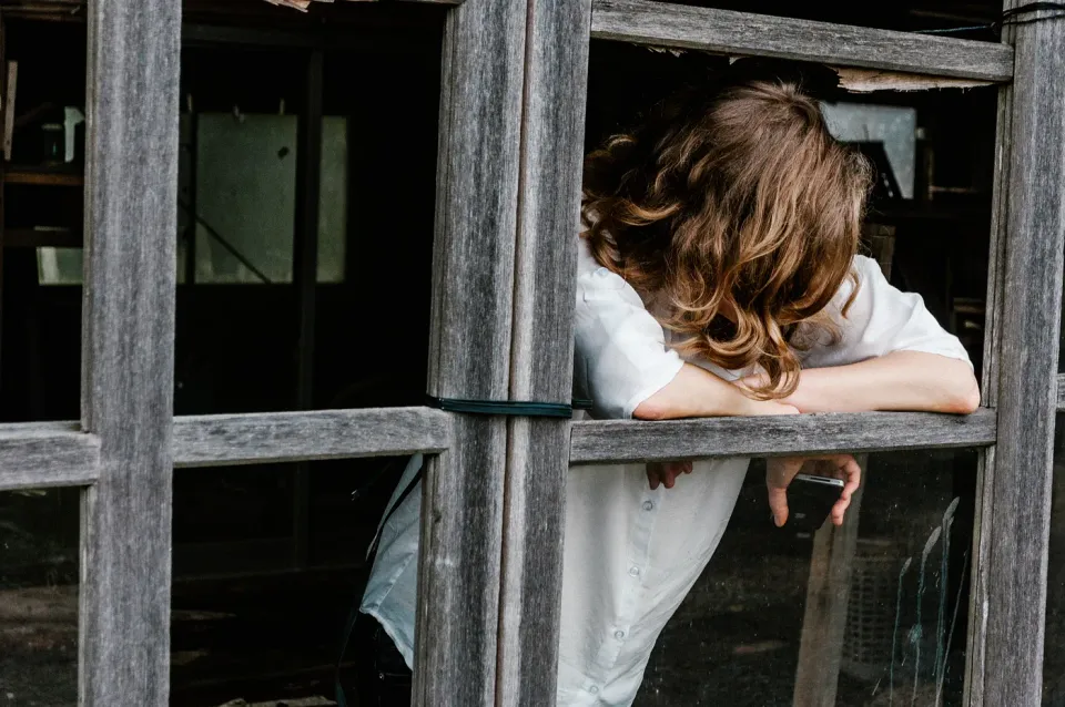 A person with medium length wavy brown hair and a short sleeve white shirt leans on a natural wood window frame