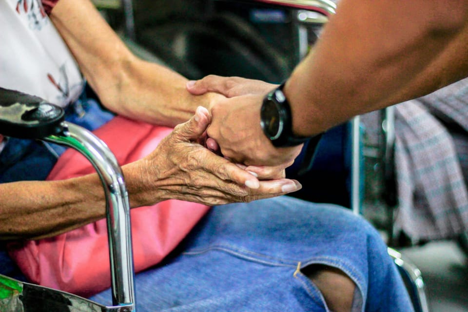 A white man's hands hold the hands of an elderly woman in a blue skirt, seated in a wheelchair