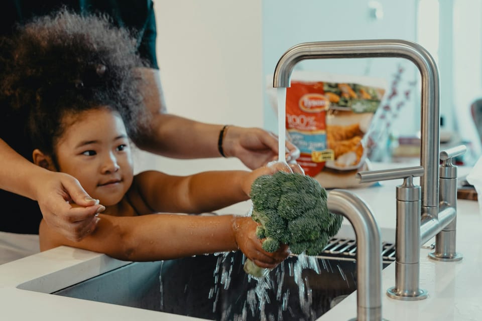 An Asian child with natural hair washes a head of broccoli under running water over a sink, an adult's hands behind her